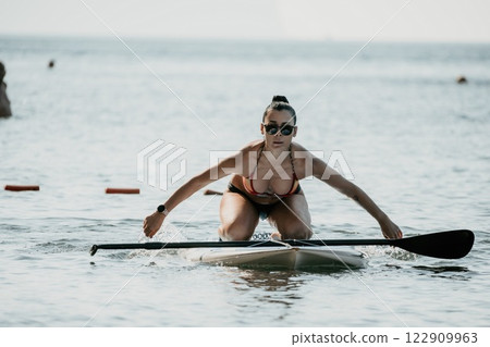 Sea woman sup. Silhouette of happy middle aged tanned woman in rainbow bikini, surfing on SUP board, confident paddling through water surface. Idyllic sunset. Active lifestyle at sea or river. 122909963