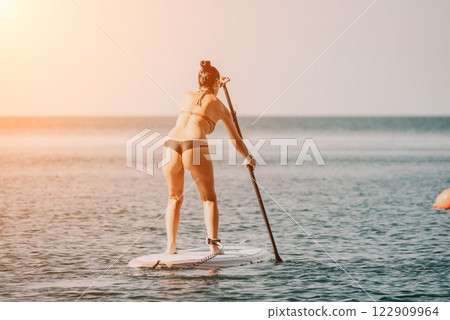 Sea woman sup. Silhouette of happy middle aged tanned woman in rainbow bikini, surfing on SUP board, confident paddling through water surface. Idyllic sunset. Active lifestyle at sea or river. 122909964