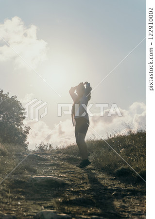 Woman Silhouette Mountain Landscape Sun A silhouette of a woman standing on a mountain with a bright sun and cloudy sky in the background. 122910002