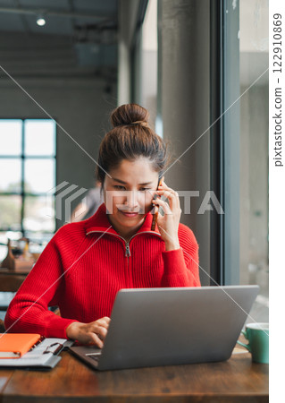 A young woman in a red sweater multitasks on her laptop while talking on the phone in a cozy cafe. A young woman in a red sweater multitasks on her laptop while talking on the phone in a cozy cafe. 122910869