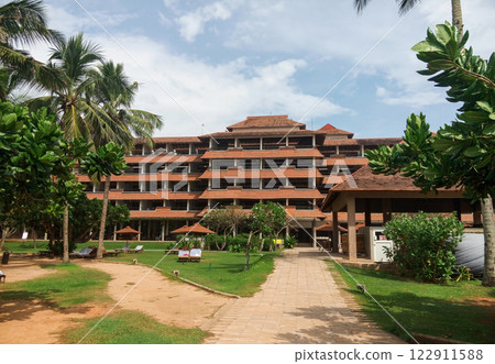 Tropical resort with a lush green garden, palm trees, and a modern multi-story building under a blue sky Tropical resort with a lush green garden, palm trees, and a modern multi-story building under a blue sky 122911588