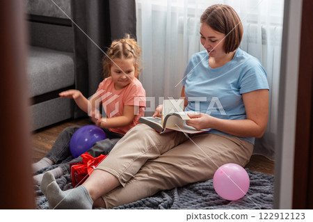 Mid shot of Caucasian little happy girl and her mother sits on carpet in room and reads book. Woman and her daughter spend time. Mother's Day Concept 122912123