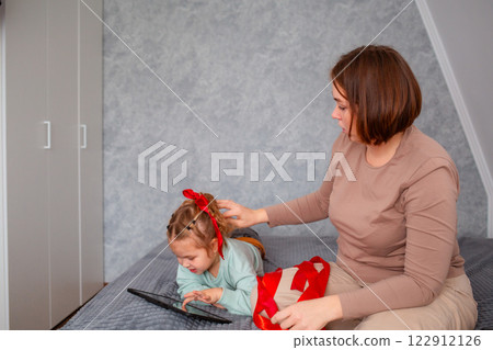 Mid shot of young Caucasian mother braids her daughter's hair while sitting on the couch. A little girl uses a tablet 122912126