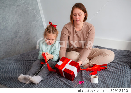 Mid shot of Caucasian mother and little daughter pack a holiday gift together. Woman teaches a child holiday preparation. Family celebration of Mother's Day and Valentine's Day 122912130