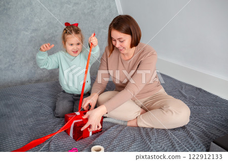 Happy Caucasian mother and little daughter pack a holiday gift together. Family celebration of Mother's Day and Valentine's Day Happy Caucasian mother and little daughter pack a holiday gift together. Family celebration of Mother's Day and Valentine's Day 122912133