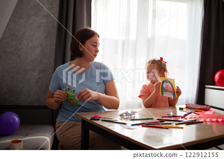 Kindergarten teacher plays and teaches cute little girl how to make paper cards for Mother's Day and Valentine's Day. Woman and child sitting at the table 122912155