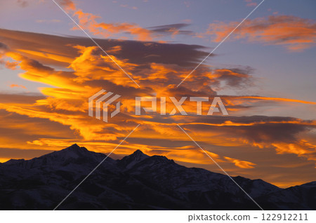 High mountains and amazing lights of a bright sunset. Thick clouds are illuminated with orange light at sunset, enveloping snow-capped mountains in shadow 122912211