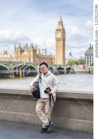 An Asian traveler holding a camera and wearing a camera bag stands near the River Thames with Big Ben and Westminster in the background. 122912596