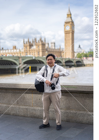 An Asian traveler holding a camera and wearing a camera bag stands near the River Thames with Big Ben and Westminster in the background. 122912602