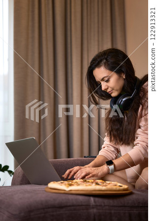 Caucasian woman working on laptop computer and eating pizza on sofa in living room 122913321