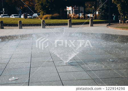 Water droplets spread from the fountain in the air. Splashing water from a fountain in the park. Vertical fountain jets in the sidewalk on the square in the city park. 122914298