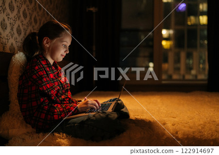 Side view of young girl sitting on bed in dark bedroom, focused using laptop looking to screen, warm lighting and soft ambiance creating cozy atmosphere. Child doing homework, enjoying digital content 122914697