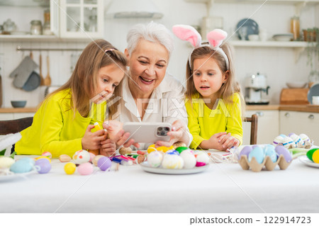 Happy Easter. Two girls sisters twins grandmother painting eggs watching video on phone enjoying time together. Happy family preparing for Easter. Good time at home. Christian festival tradition Happy Easter. Two girls sisters twins grandmother painting eggs watching video on phone enjoying time together. Happy family preparing for Easter. Good time at home. Christian festival tradition 122914723