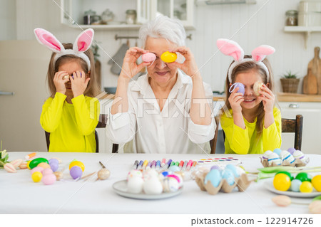 Happy Easter. Two little girls sisters twins grandmother painting eggs enjoying time together. Happy family grandma granddaughters child kids preparing for Easter. Spring Christian festival tradition Happy Easter. Two little girls sisters twins grandmother painting eggs enjoying time together. Happy family grandma granddaughters child kids preparing for Easter. Spring Christian festival tradition 122914726