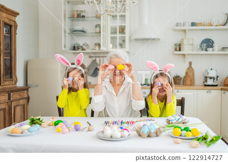 Happy Easter. Two little girls sisters twins grandmother painting eggs enjoying time together. Happy family grandma granddaughters child kids preparing for Easter. Spring Christian festival tradition 122914727