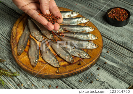 A hand sprinkles a mix of spices over neatly arranged dried fish on a wooden cutting board. The rustic kitchen setup features cracked wood and a touch of culinary artistry 122914760