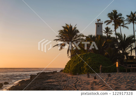 A serene beach at sunset featuring a white lighthouse, lush greenery, tall palm trees, rocky shoreline, and a calm ocean under golden light. 122917328