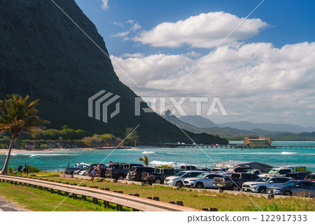 A turquoise ocean with a pier and small structure, a mountain on the left, a parking area with cars, and a palm tree under a partly cloudy sky. 122917333