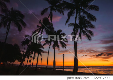 A vibrant sunset on a Hawaiian beach with silhouetted palm trees, a calm ocean, and a surfboard shaped monument near lampposts along the shore. A vibrant sunset on a Hawaiian beach with silhouetted palm trees, a calm ocean, and a surfboard shaped monument near lampposts along the shore. 122917352