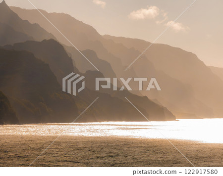 The Napali Coast on Kauai Island features jagged cliffs in golden light, silhouetted against the sky with the ocean reflecting the warm hues below. The Napali Coast on Kauai Island features jagged cliffs in golden light, silhouetted against the sky with the ocean reflecting the warm hues below. 122917580