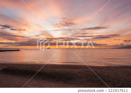 A serene sunset over calm ocean waters at Waikiki Beach, Oahu. Vibrant sky hues, sandy beach, grass, boats, and a pier create a tranquil scene. A serene sunset over calm ocean waters at Waikiki Beach, Oahu. Vibrant sky hues, sandy beach, grass, boats, and a pier create a tranquil scene. 122917611