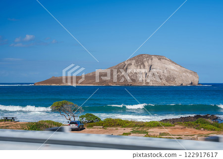 View of Rabbit Island's dome like shape off Oahu's coast, with a coastal park featuring a picnic table, parked car, and rocky shoreline. 122917617