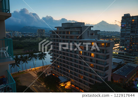 A cityscape of Honolulu at dusk featuring a lit multi story building, a canal lined with palm trees, and mountains silhouetted against the sky. 122917644