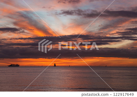 Vibrant sunset with orange, pink, and purple hues over calm ocean waters. A cruise ship and sailboats, including one with a triangular sail, are visible. 122917649