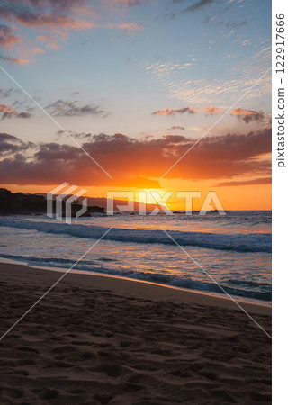 Golden orange sunset illuminates the sky and ocean waves, with soft sandy shoreline, visible footprints, and rocky outcroppings in Hawaii. 122917666