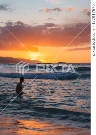 A vibrant sunset casts orange and pink hues over calm ocean waters. A lone individual wades in the waves, with a distant landmass silhouetted in the background. 122917670
