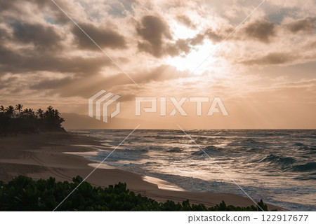 A serene Hawaiian beach at sunset with golden light over ocean waves, scattered clouds, lush greenery, palm trees, and distant hills. A serene Hawaiian beach at sunset with golden light over ocean waves, scattered clouds, lush greenery, palm trees, and distant hills. 122917677