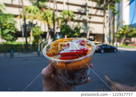 A colorful acai bowl with strawberries, mango, coconut, and granola is held against a modern building, palm trees, and a street in Honolulu. A colorful acai bowl with strawberries, mango, coconut, and granola is held against a modern building, palm trees, and a street in Honolulu. 122917757