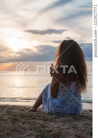 A woman with wavy hair sits on a sandy beach during sunset, facing the ocean. The sky glows with orange and blue hues, with boats visible in the distance. 122917771
