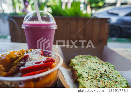 A colorful acai bowl with fruit, avocado toast, and a pink smoothie on a table. Blurred greenery and a parked car suggest a tropical setting. 122917784
