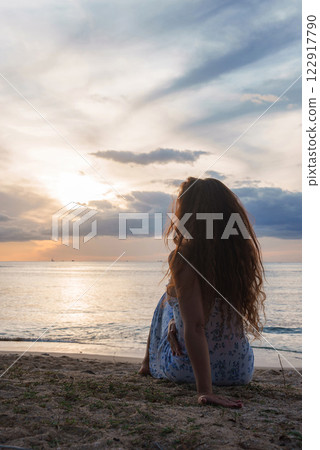 A woman with wavy hair sits on a beach, facing a calm ocean at sunset. The sky features orange and blue hues, with boats visible on the horizon. A woman with wavy hair sits on a beach, facing a calm ocean at sunset. The sky features orange and blue hues, with boats visible on the horizon. 122917790