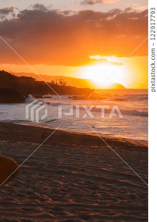 A warm orange sunset reflects on ocean waves near a sandy Hawaiian beach. A surfboard rests on the shore, with cliffs and trees in the distance. 122917793