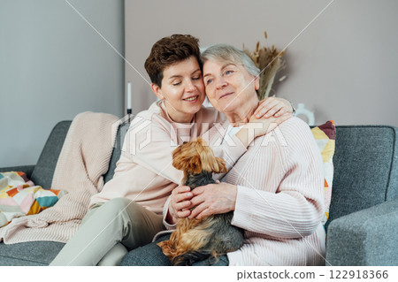 Smiling senior 70s woman with daughter, Yorkshire terriers dog pet hugging on sofa. Family weekend together. Parenting love, care, generations unity concept. Spending quality time in retiring house. 122918366