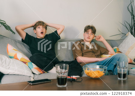 Two excited teenager boys watching match game together at home. Brothers seated on couch with snacks and drinks, cheering for favourite team but are Disappointed after Team Loses. Young sport fans 122918374