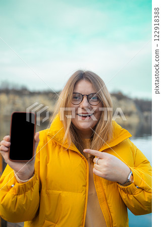 Beautiful Caucasian woman in yellow jacket stands outdoor and shows her smartphone to the camera. the smartphone with a black screen mockup 122918388