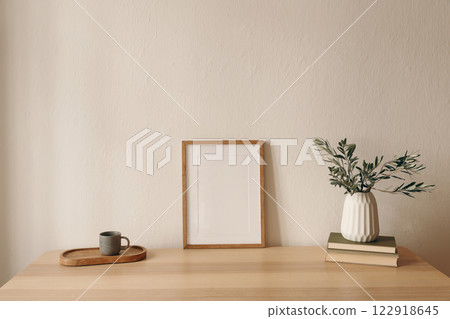 Summer interior still life. Blank wooden picture frame mockup. Modern interior, minimal scandinavian home design. Olive tree branches in vase, cup of coffee tea, books on table. Beige wall background. Summer interior still life. Blank wooden picture frame mockup. Modern interior, minimal scandinavian home design. Olive tree branches in vase, cup of coffee tea, books on table. Beige wall background. 122918645