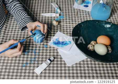 Young boys kids decorating Easter eggs. Closeup of childrens hands with paint brush, acrylic paint tube. Birds eggs in ceramic bowl. Checkered table cloth. Blurred background. Craft creative concept. Young boys kids decorating Easter eggs. Closeup of childrens hands with paint brush, acrylic paint tube. Birds eggs in ceramic bowl. Checkered table cloth. Blurred background. Craft creative concept. 122919125