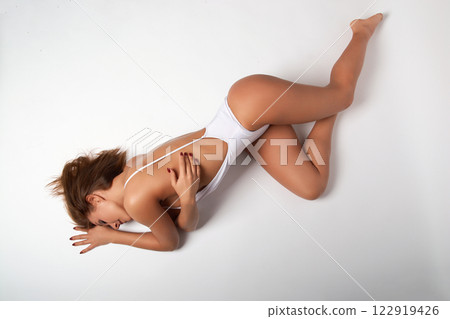 A girl in a fitted white bodysuit lies on a perfectly even white background. 122919426