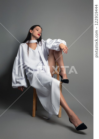 A brunette girl in a white dress is sitting on a chair on a gray background 122919444