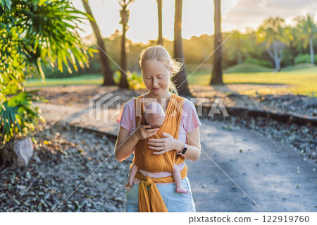 A loving mother walks through a peaceful park, carrying her baby securely in a sling. A heartwarming moment of maternal bonding, babywearing, and the joy of motherhood in nature 122919760