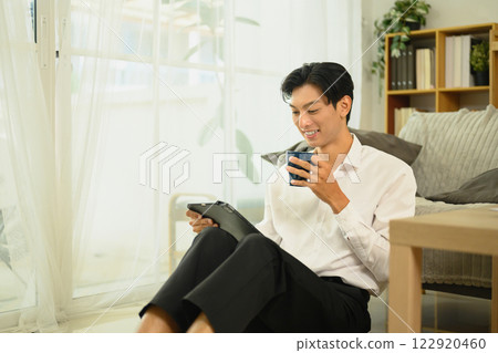 Smiling young man sitting near a window sipping coffee and using digital tablet Smiling young man sitting near a window sipping coffee and using digital tablet 122920460