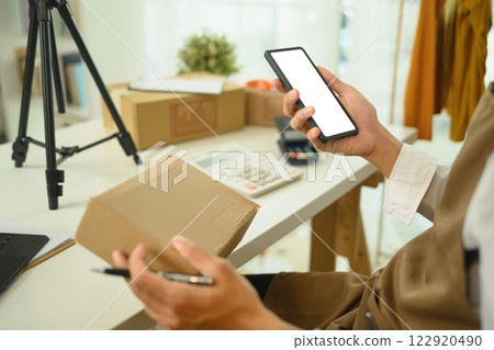 Close up of an entrepreneur checking orders on smartphone at a workspace filled with shipping boxes 122920490