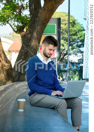 Smiling male freelancer sitting on curved bench under a tree and working on his laptop Smiling male freelancer sitting on curved bench under a tree and working on his laptop 122920552