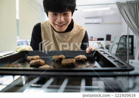 Cheerful asian man putting tray with chocolate chip cookies into oven 122920616