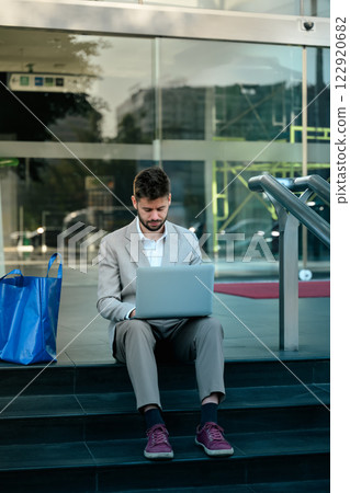 Professional businessman sitting on the steps outside a modern glass building, working on his laptop Professional businessman sitting on the steps outside a modern glass building, working on his laptop 122920682