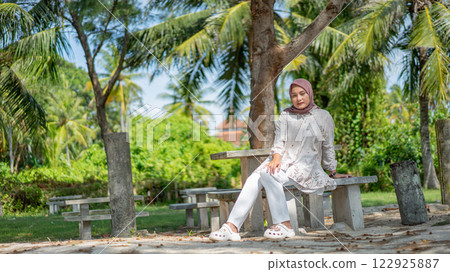 Woman in Hijab Relaxing on a Bench Under Palm Trees at a Tropical Beach Resort 122925887